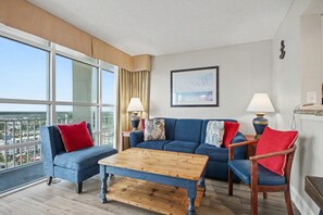 A modern living room with a large window, a blue sofa set, a wooden coffee table, and a lamp. - Oceanview 1BR Suite + Lazy River & Hot Tub (Myrtle Beach)