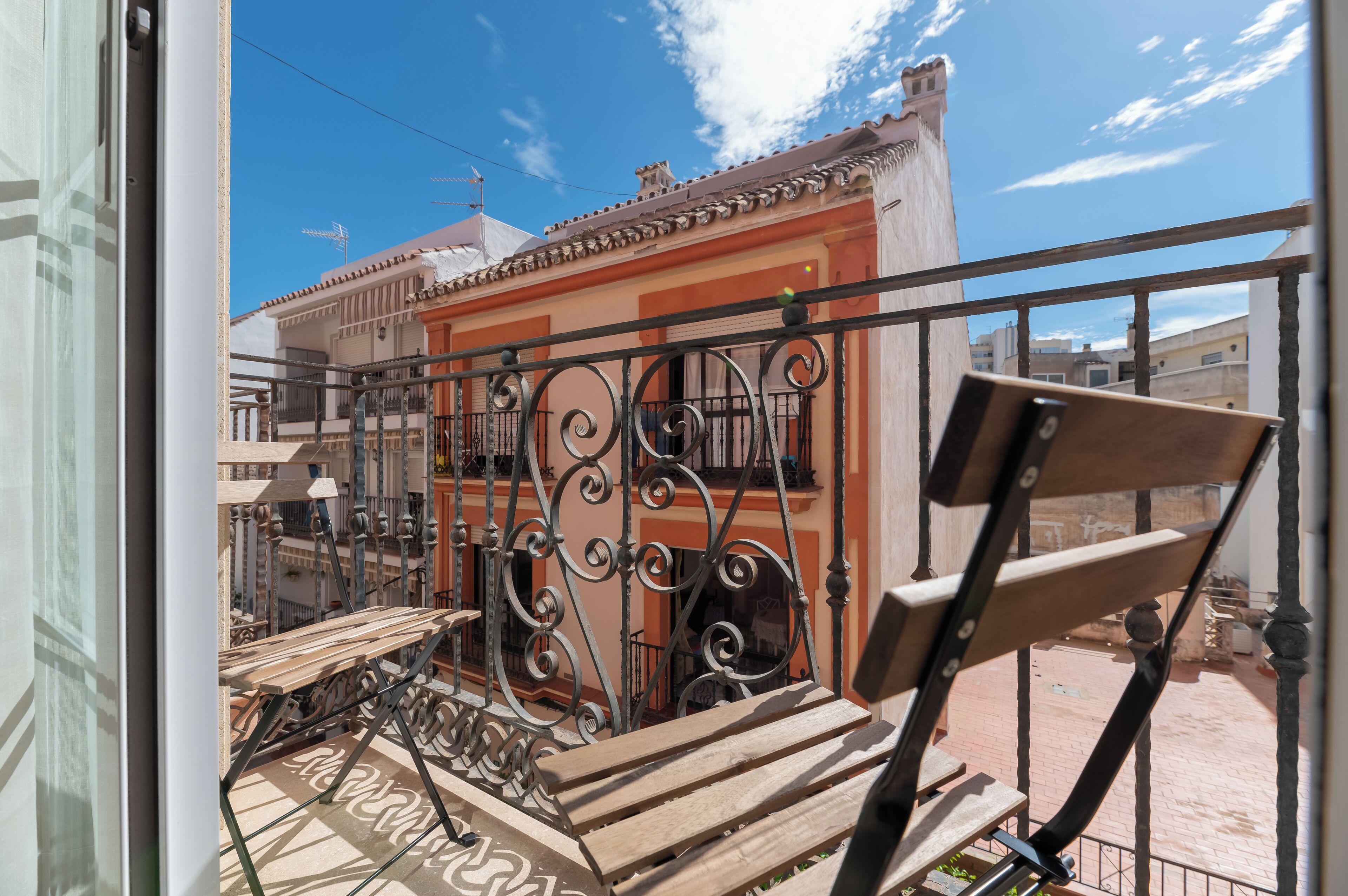A balcony with a wrought iron railing, a wooden bench, and a view of a building with a tiled roof.