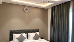 A modern hotel room with a tufted headboard, a bed with white linens and grey pillows, a round wall clock, and a large window with dark curtains. - LG CORDIAL (Mysore)