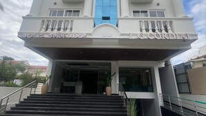 A multi-story hotel with a central blue glass facade, a red carpeted entrance, and a modern architectural design. - LG CORDIAL (Mysore)