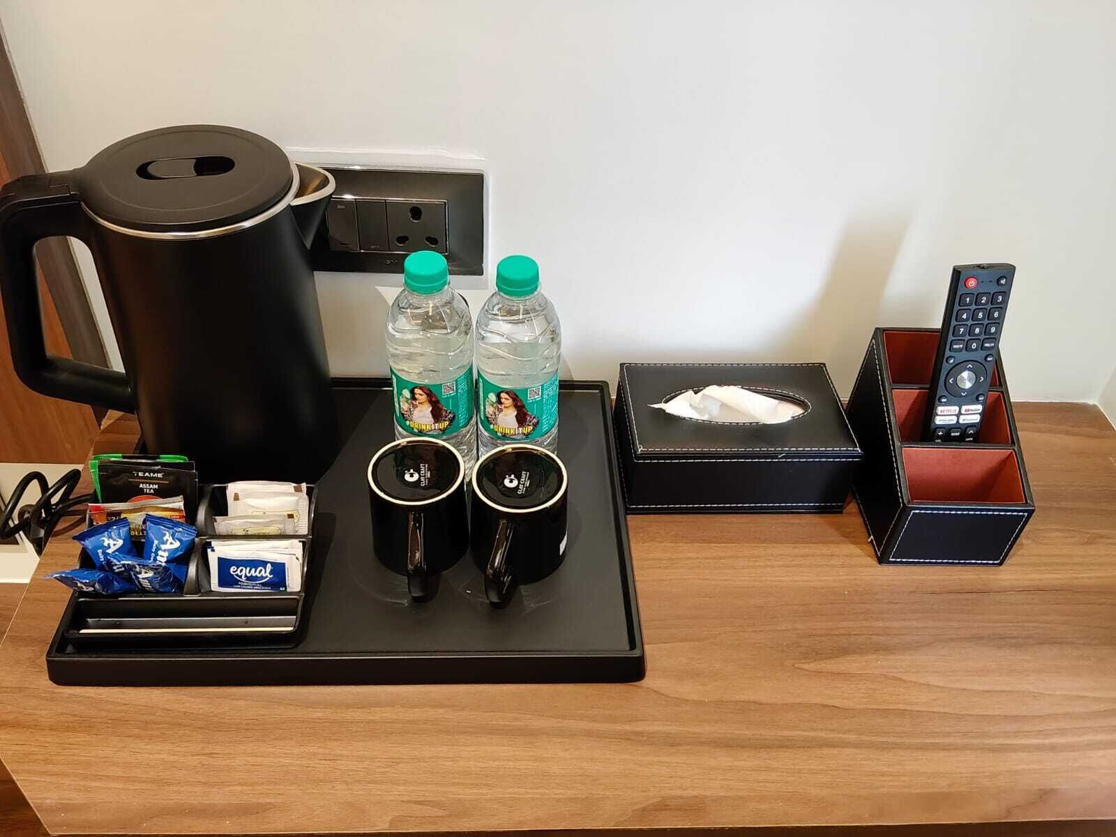 A hotel room desk with a coffee maker, two water bottles, a tissue box, a remote control, and a tray with tea bags and snacks.