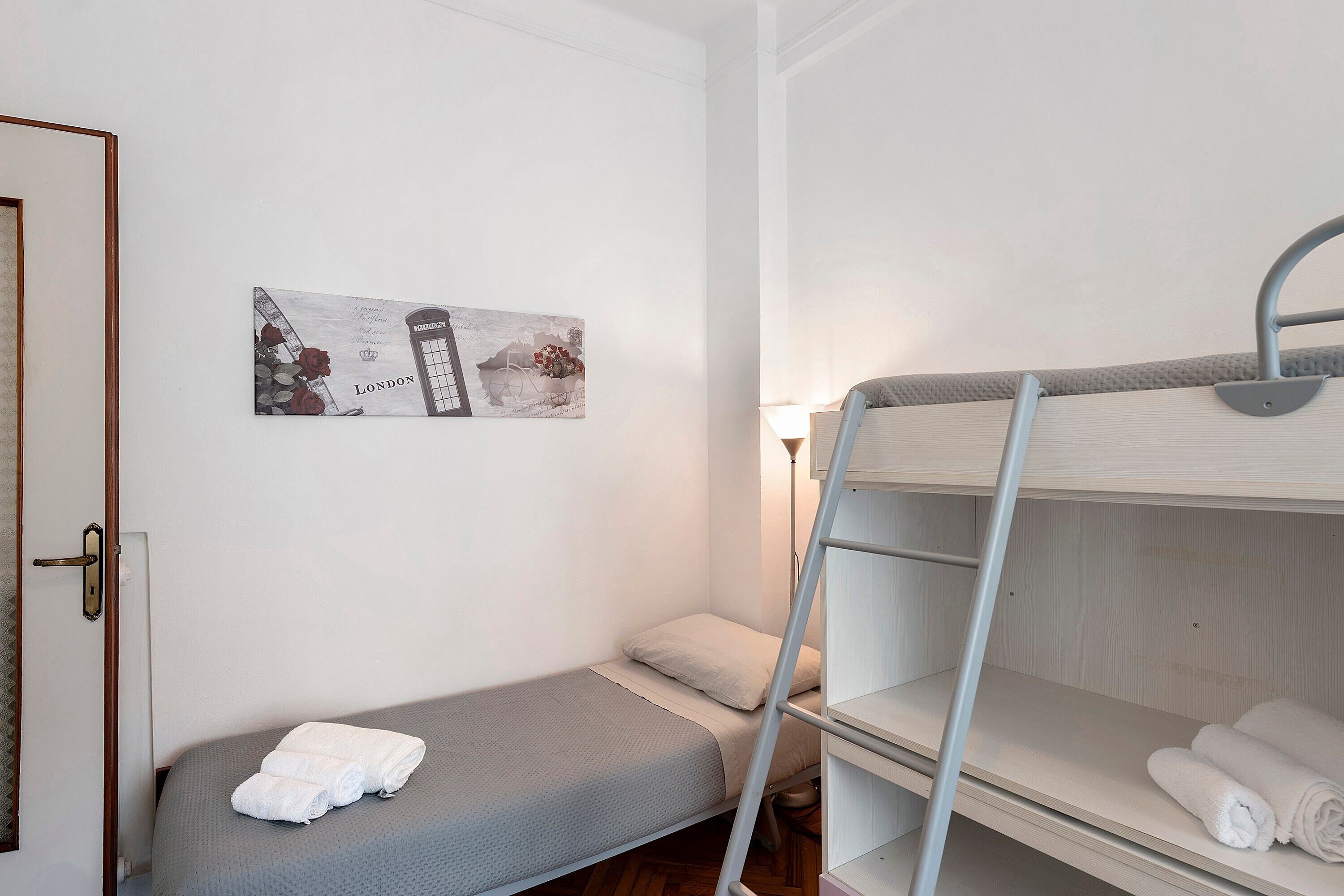 Een enkel bed met een grijze matras en witte lakens, een stapelbed met witte lakens, een lijst met een foto van Londen en een deur met een knop.