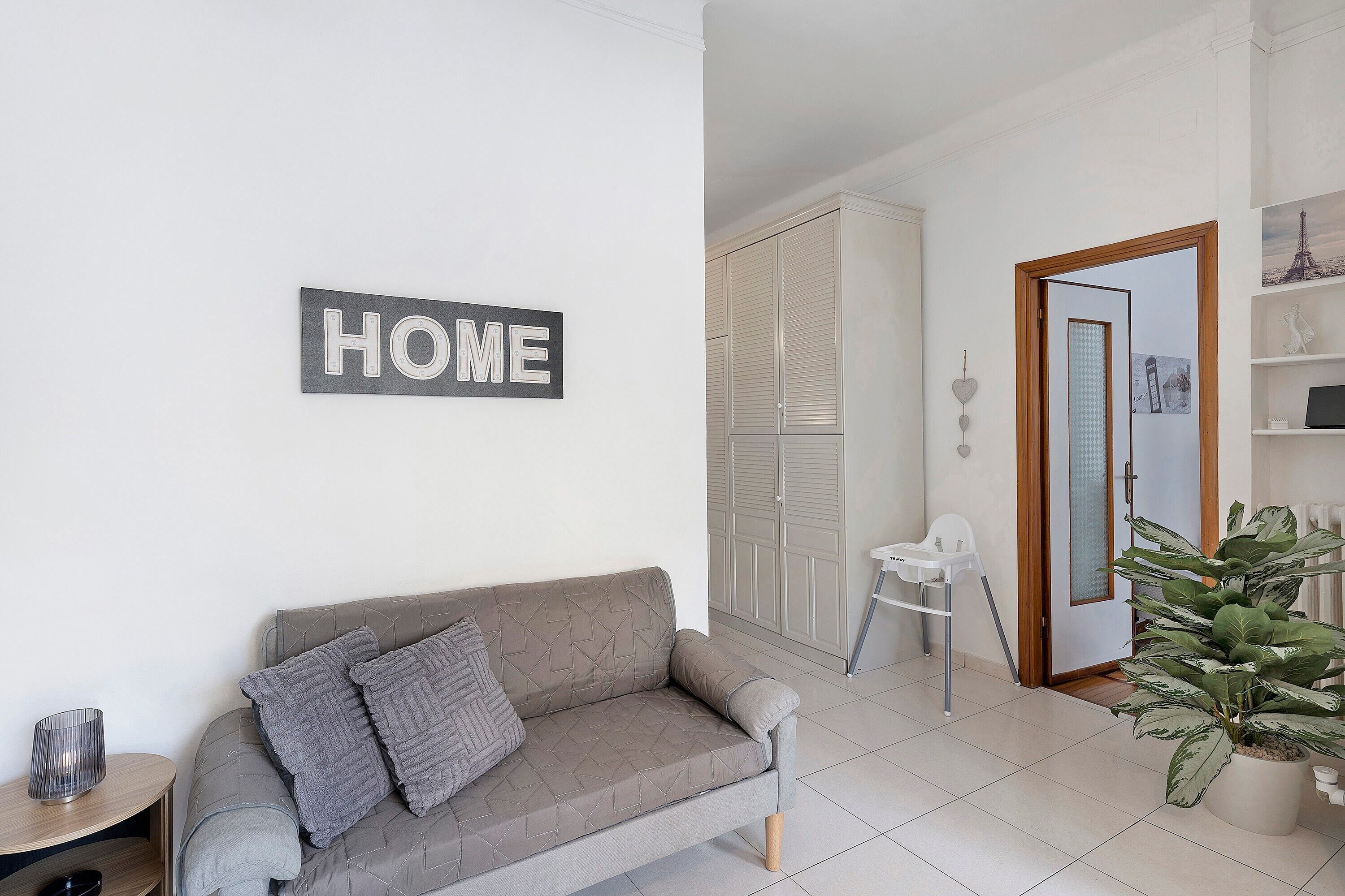 A neutral-toned living room with a sofa, a 'HOME' sign, a potted plant, and a white wardrobe.