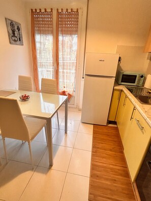 Eine kleine Küche mit Kühlschrank, Mikrowelle und Spüle. Ein Esstisch mit Stühlen und ein Fenster mit Vorhängen sind ebenfalls vorhanden.