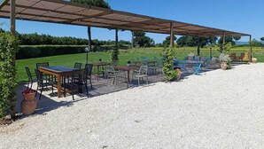 Patio esterno con tavoli e sedie, un pergolato e terreno sterrato.