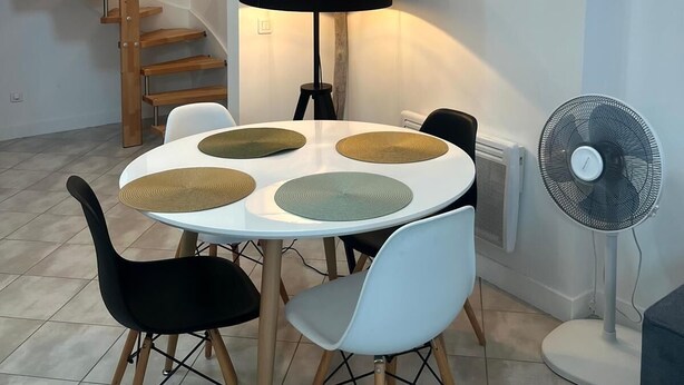 A dining area with a round table, four chairs, a black lamp, a hanging light fixture, a wall-mounted light, and a standing fan. - La Loge de l'Opéra – Elegant One-Bedroom Apartment in the Heart of the City (Toulon)