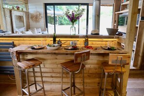 Un coin bar en bois, avec des tabourets, une grande fenĂȘtre et une vue sur la verdure environnante.