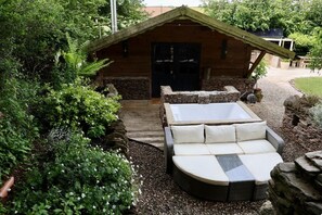 A wooden cabin with a stone exterior, a large outdoor seating area with cushions, and a gravel path leading to the entrance.