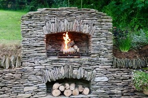 A stone-built outdoor fireplace with a fire burning, surrounded by a garden.