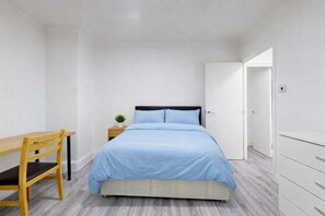 Ein Schlafzimmer mit einem Bett, einem Schreibtisch, einem Stuhl, einem Nachttisch mit Pflanze und einem Kleiderschrank.