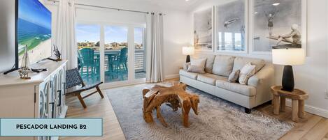A spacious living room with a large window, a ceiling fan, a wooden coffee table, and a sofa with a view of the ocean.