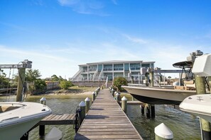Une jetée en bois mène à un bâtiment moderne à plusieurs niveaux, doté de balcons, et entouré d’eau et de bateaux.
