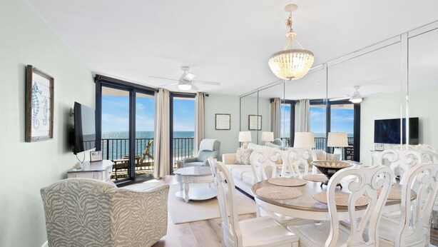 Un salón espacioso con un ventanal grande con vistas al mar, una araña y una zona de comedor con mobiliario blanco.