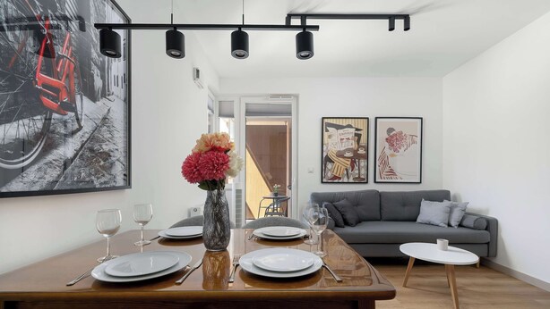 Un comedor con una mesa de madera dispuesta para cuatro, un jarrón con flores y un sofá gris al fondo.