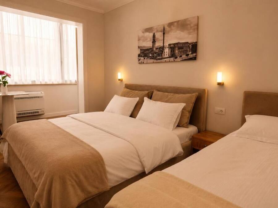 A hotel room with a large bed, a smaller bed, a desk with a chair, a window with curtains, and a wall-mounted picture.