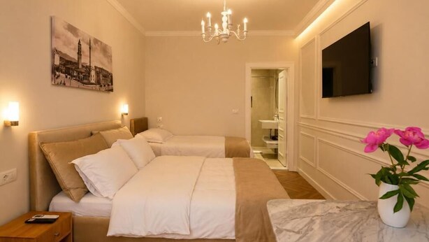 A hotel room with two beds, a chandelier, a flat-screen TV, a painting, and a vase with flowers.