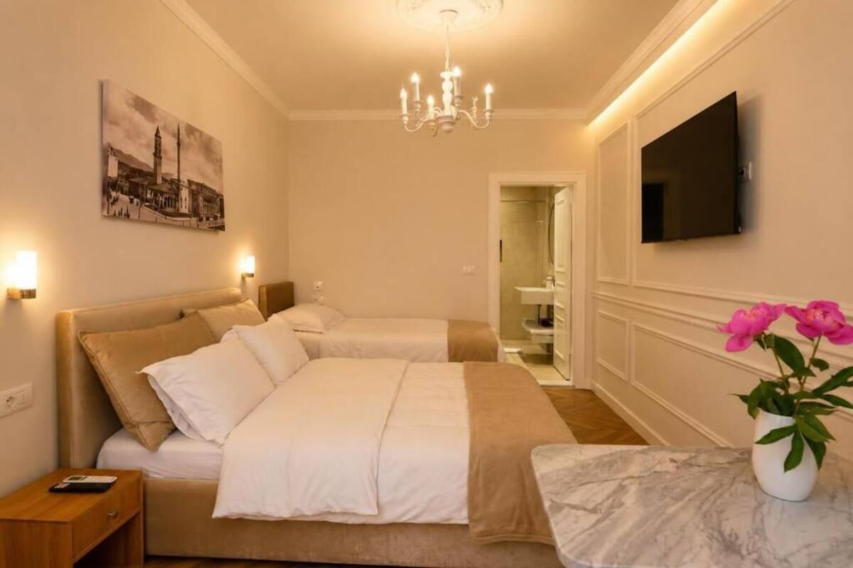 A hotel room with two beds, a chandelier, a flat-screen TV, a painting, and a vase with flowers.