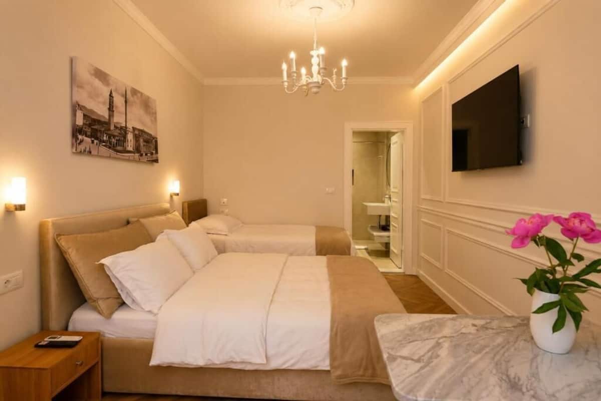 A hotel room with two beds, a chandelier, a flat-screen TV, a painting, and a vase with flowers.
