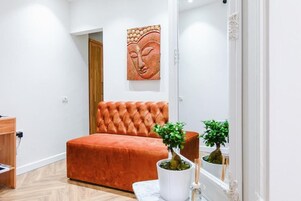 Un dormitorio con un sofá naranja capitonado, una puerta de madera, una obra de arte colgada en la pared y dos plantas en macetas.