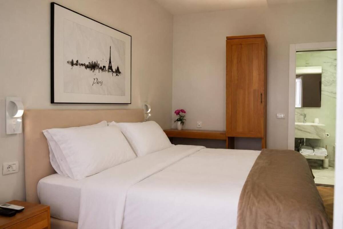 A bedroom with a bed, a wooden wardrobe, a nightstand, a framed picture of Paris, and a bathroom visible through an open door.