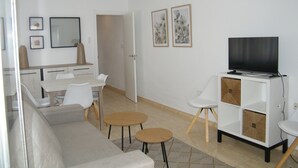 A modern living room with a sofa, a TV on a cabinet, a dining table with chairs, and a kitchen area in the background. - Family apartment, sea view, parking - 7 guests (Gandia)