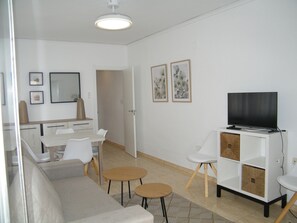 Ein modernes Wohnzimmer mit Sofa, Fernseher auf einer Kommode, Esstisch mit Stühlen und einer Küche im Hintergrund.