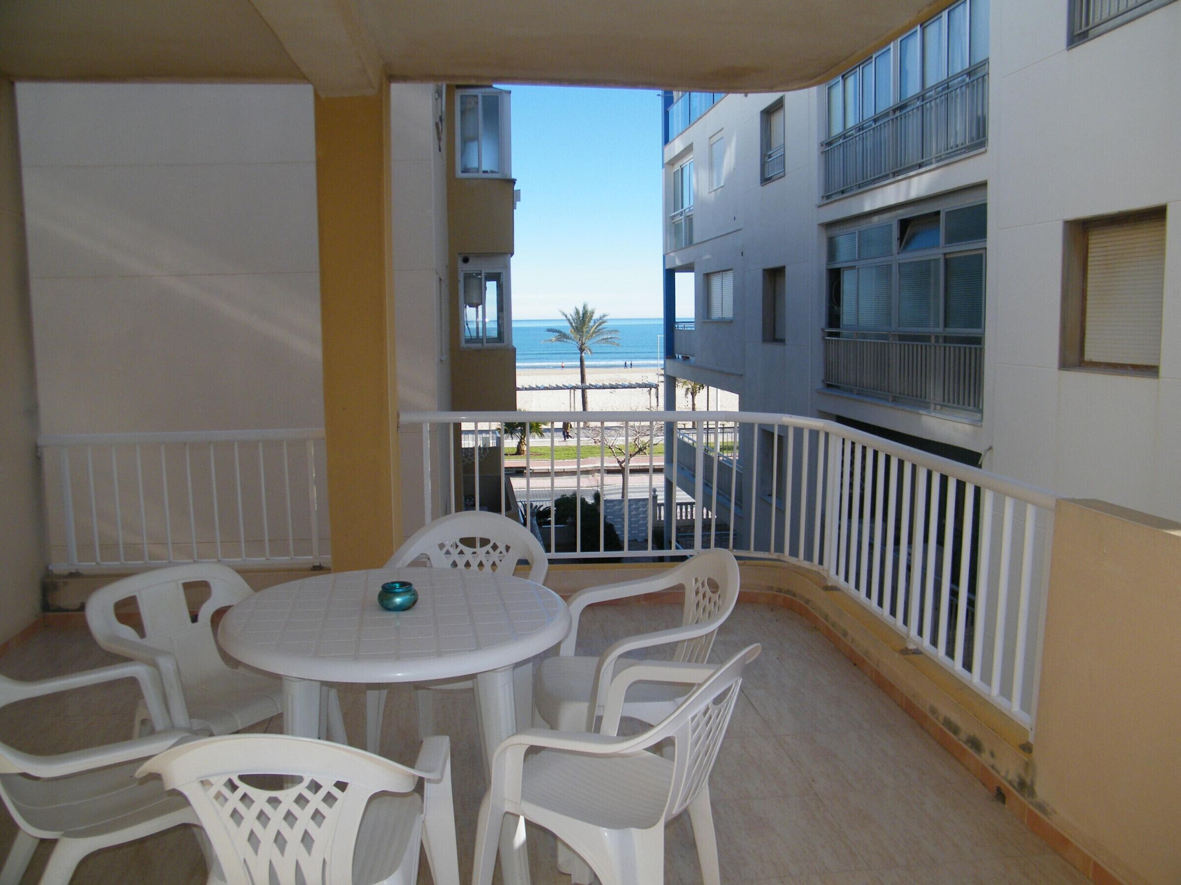 A balcony with a white table and chairs, overlooking a beach and buildings.