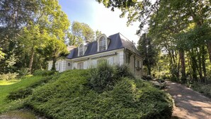 Une grande maison à deux étages, dotée d’un toit à forte pente, entourée d’une végétation luxuriante et d’arbres.
