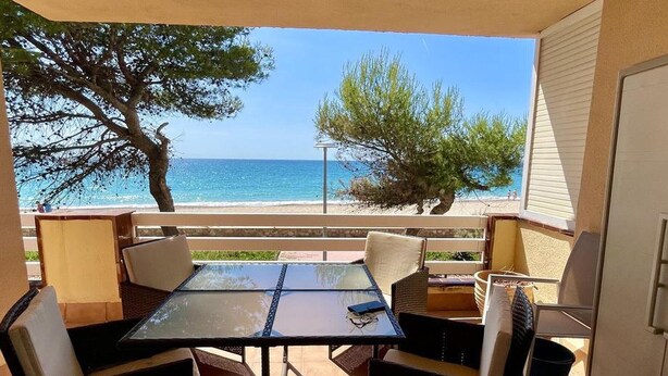 A balcony with a glass table and chairs, overlooking a beach and the sea.