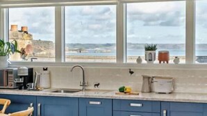A kitchen with a large window overlooking a beach, a sink, and various kitchen items on the counter. - Sea-View Apartment - Steps to Beach & Shops (Pembrokeshire)