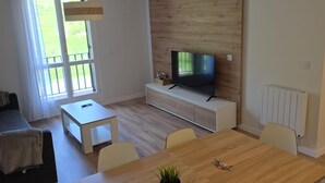 A modern living room with a TV on a wooden wall, a dining table with chairs, a sofa, and a window with curtains. - Modern studio near slopes - 4 guests (Aisa-Candanchú)