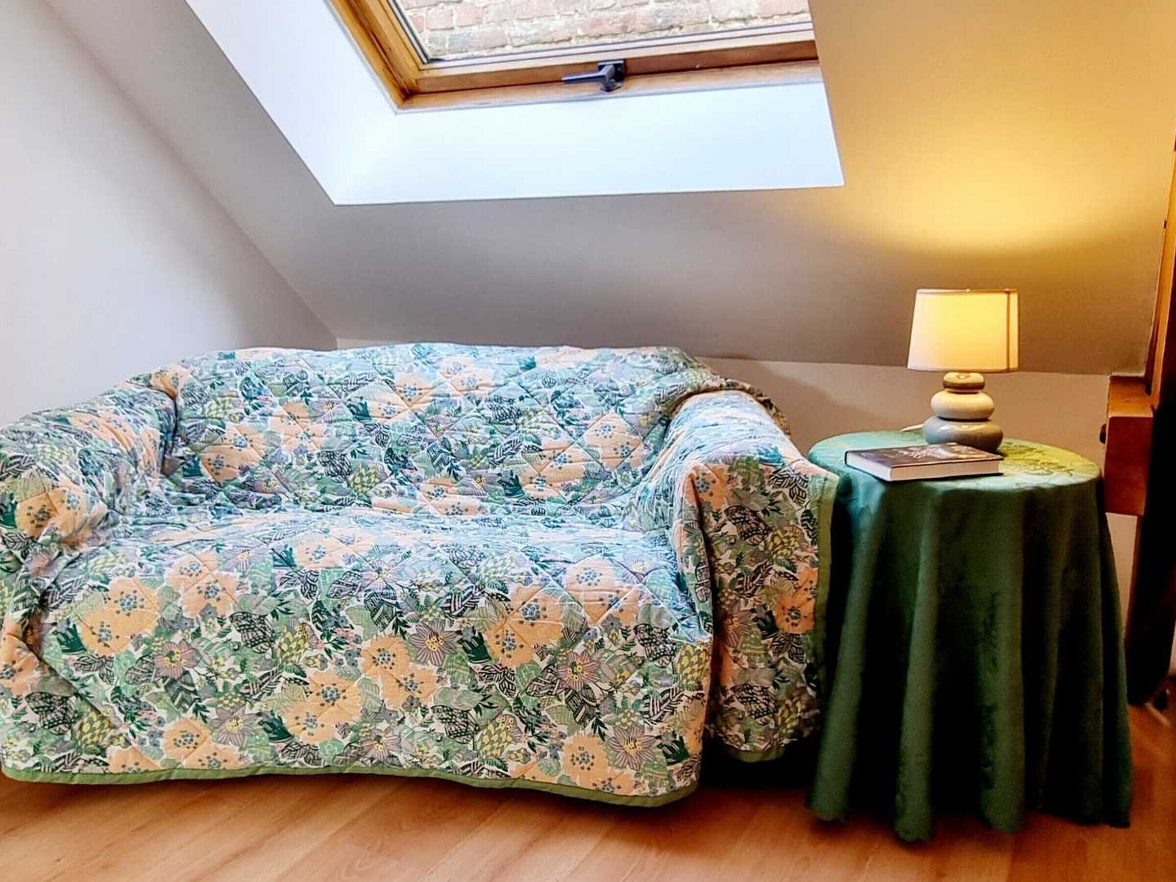 A room with a skylight, a floral-patterned sofa, a small table with a lamp, and a book.