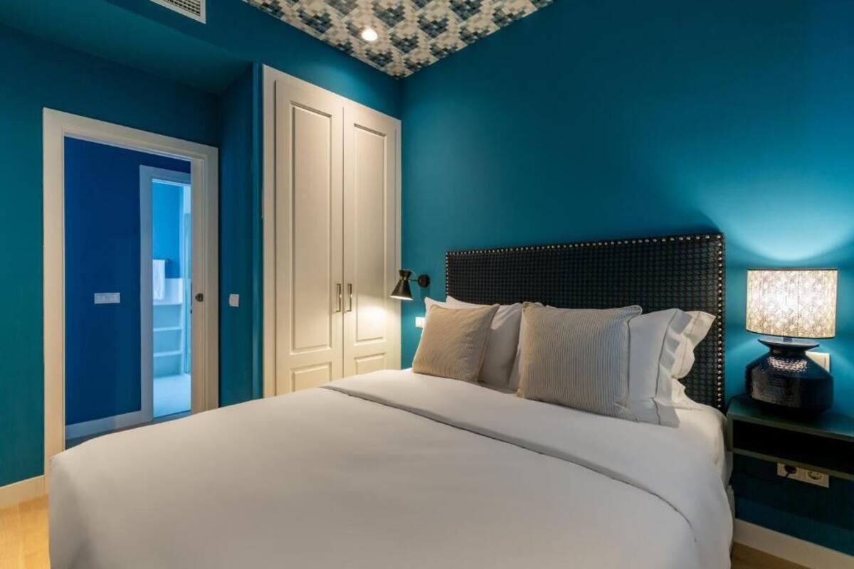 A bedroom with a large bed, a headboard, a nightstand with a lamp, and a door leading to a bathroom.