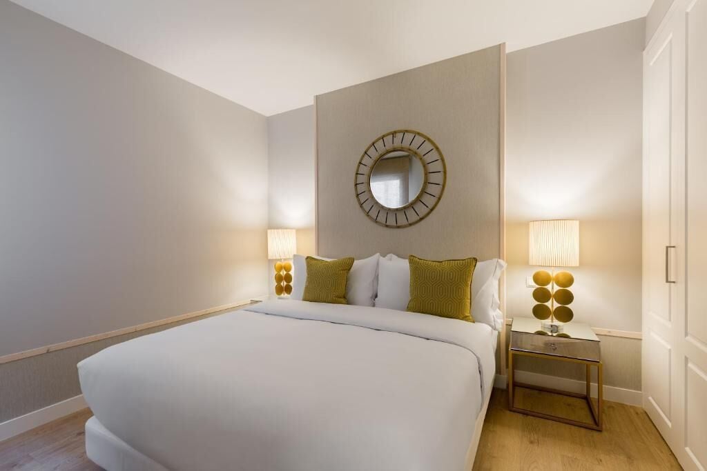 Ein Hotelzimmer mit einem großen Bett, zwei Nachttischen mit Lampen, einem runden Spiegel an der Wand und einer Tür rechts.