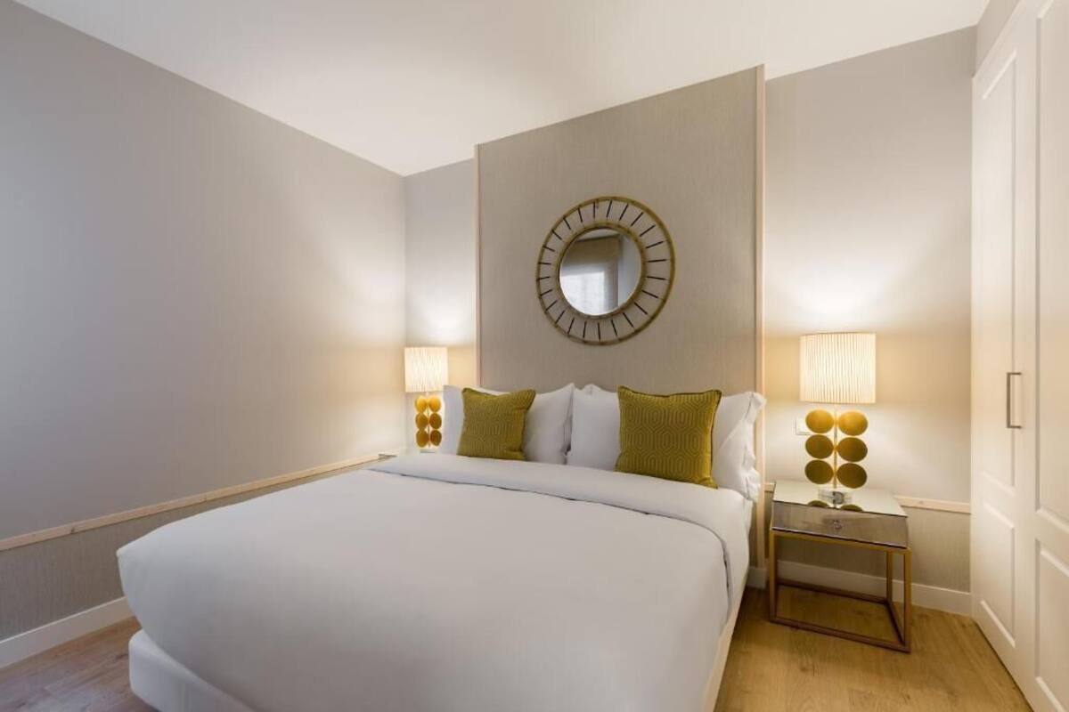 A hotel room with a large bed, two bedside tables with lamps, a round mirror on the wall, and a door on the right.
