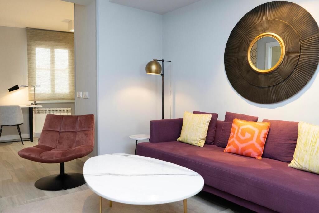 Una sala de estar moderna con un sofá morado, un espejo redondo, una lámpara de pie y una silla roja.