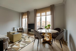 Ein modernes Wohnzimmer mit einer Couch, einem Couchtisch, einem Esstisch mit Stühlen, einem Fernseher und großen Fenstern mit Vorhängen.