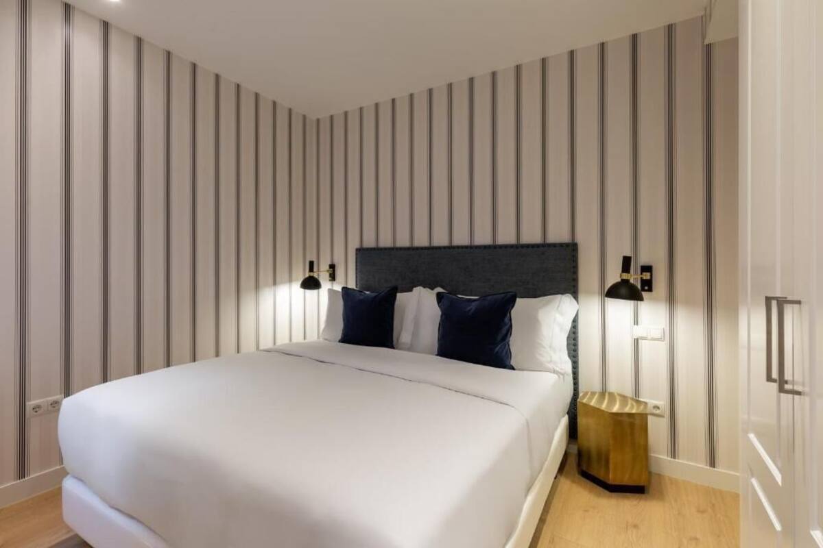 A modern hotel room with a large bed, striped wallpaper, and a small bedside table with a lamp.
