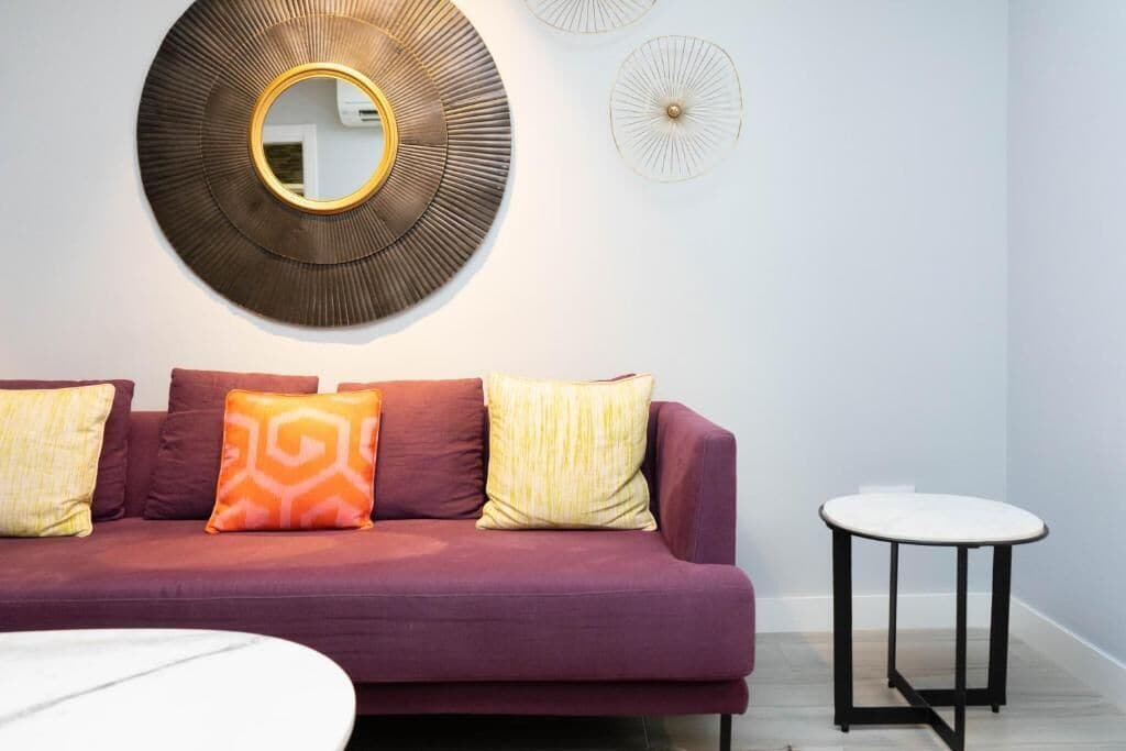 A modern living room with a purple sofa, patterned cushions, a round mirror, and a small round table.