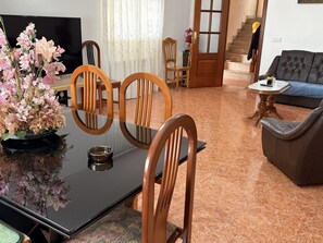 Een eethoek met een tafel, stoelen, een bloevas, een televisie en een bank op de achtergrond.