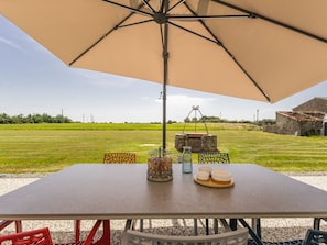 A table with an umbrella, outdoor seating, a decorative metal structure, and a view of a green field and stone building.