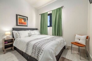 Quarto com cama, mesa de cabeceira, cadeira, janela com cortinas e um quadro na parede.