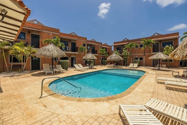Uma piscina com espreguiçadeiras e guarda-sóis de palha, rodeada por edifícios de tijolo com varandas.
