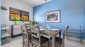 Zona pranzo con tavolo apparecchiato per quattro, sedie, ventilatore a soffitto e parete blu con un quadro incorniciato.