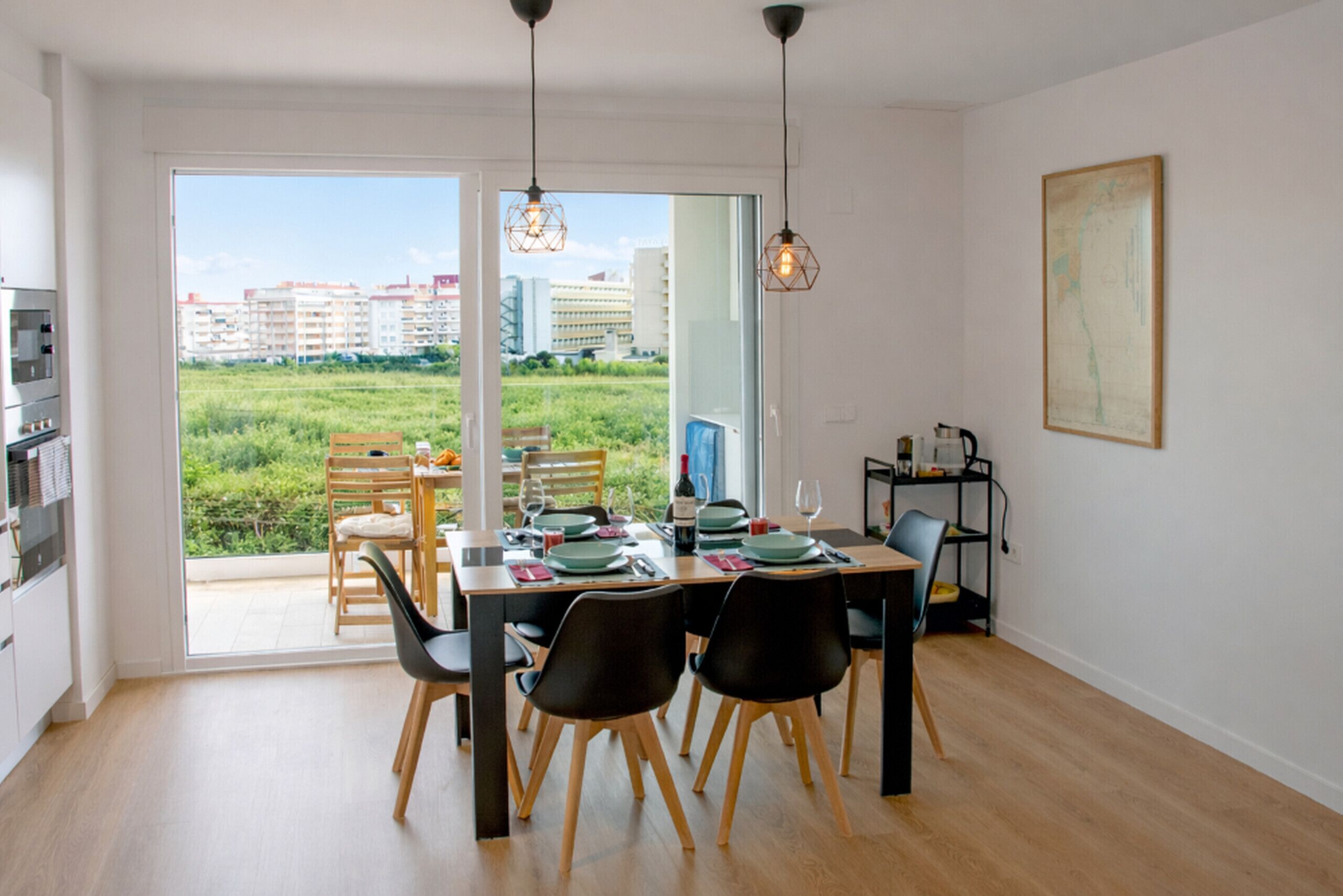 A dining area with a table set for four, a view of greenery and buildings through a glass door, and a painting on the wall. - Apartamento La Mar Inmomart (Peñiscola)