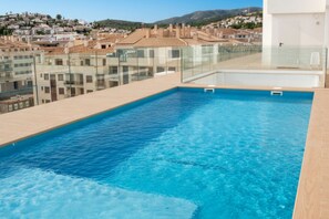 A rooftop pool with clear blue water, surrounded by a glass balustrade, overlooking a cityscape with buildings and hills. - Apartamento La Mar Inmomart (Peñiscola)