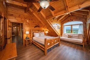 Ein Schlafzimmer in einer Holzhütte mit einem Bett, einer Couch, einem Deckenventilator und einem großen Fenster.