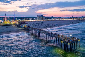 Eine Pier, die sich ins Wasser erstreckt, mit Menschen, die darauf entlanggehen, ein Strand im Hintergrund und ein Gebäude mit Kuppeldach.