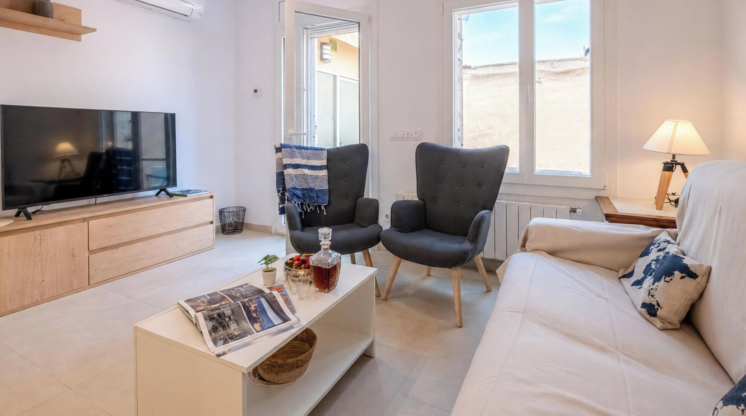 A modern living room with a flat-screen TV, two armchairs, a sofa, a coffee table with magazines, and a lamp.