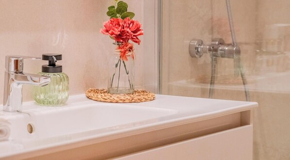 A bathroom with a glass shower enclosure, a white sink with a soap dispenser, and a decorative glass vase with flowers. - Charming house in the heart of Costa Brava (Palamós)